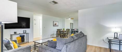 A modern living room with a grey sectional sofa, a dining area with a table and chairs, and a flat-screen TV mounted on the wall.