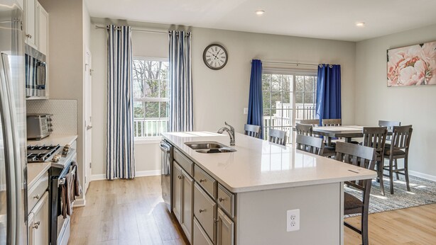 A modern kitchen with a central island, a dining area with wooden chairs, and a large window with curtains. - 30 Mi to Downtown DC: Charming Dale City Townhome! (Woodbridge)