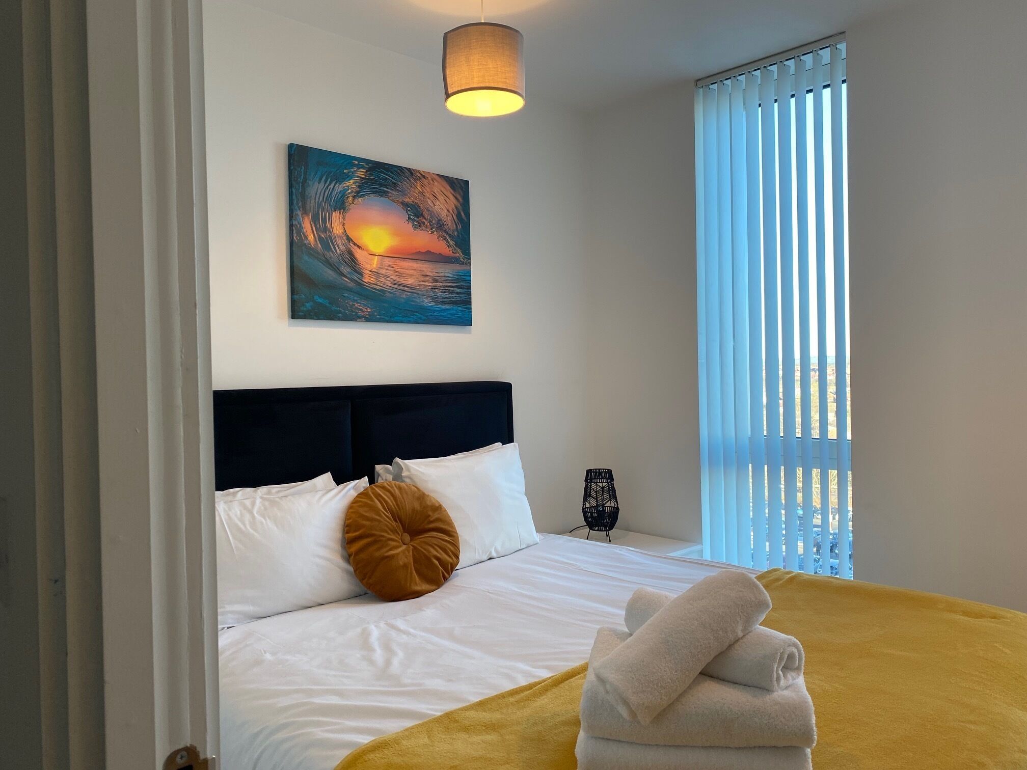 Un lit bien fait, avec du linge de lit blanc, un coussin décoratif marron et un plaid jaune. Au-dessus du lit, on peut voir une peinture représentant un coucher de soleil sur l’eau.