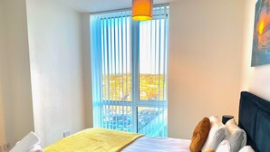 Quarto de hotel com cama, manta amarela, toalhas brancas e vista para o horizonte urbano pela janela.