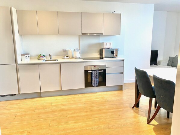 A modern kitchen with a microwave, toaster, and kettle. There is a sink and a dishwasher. The room has wooden flooring and grey chairs.