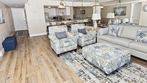 Una sala de estar moderna con un sofá, dos sillones y un reposapiés a juego, todos con estampado floral. Se observa un comedor en el fondo, junto a una cocina.