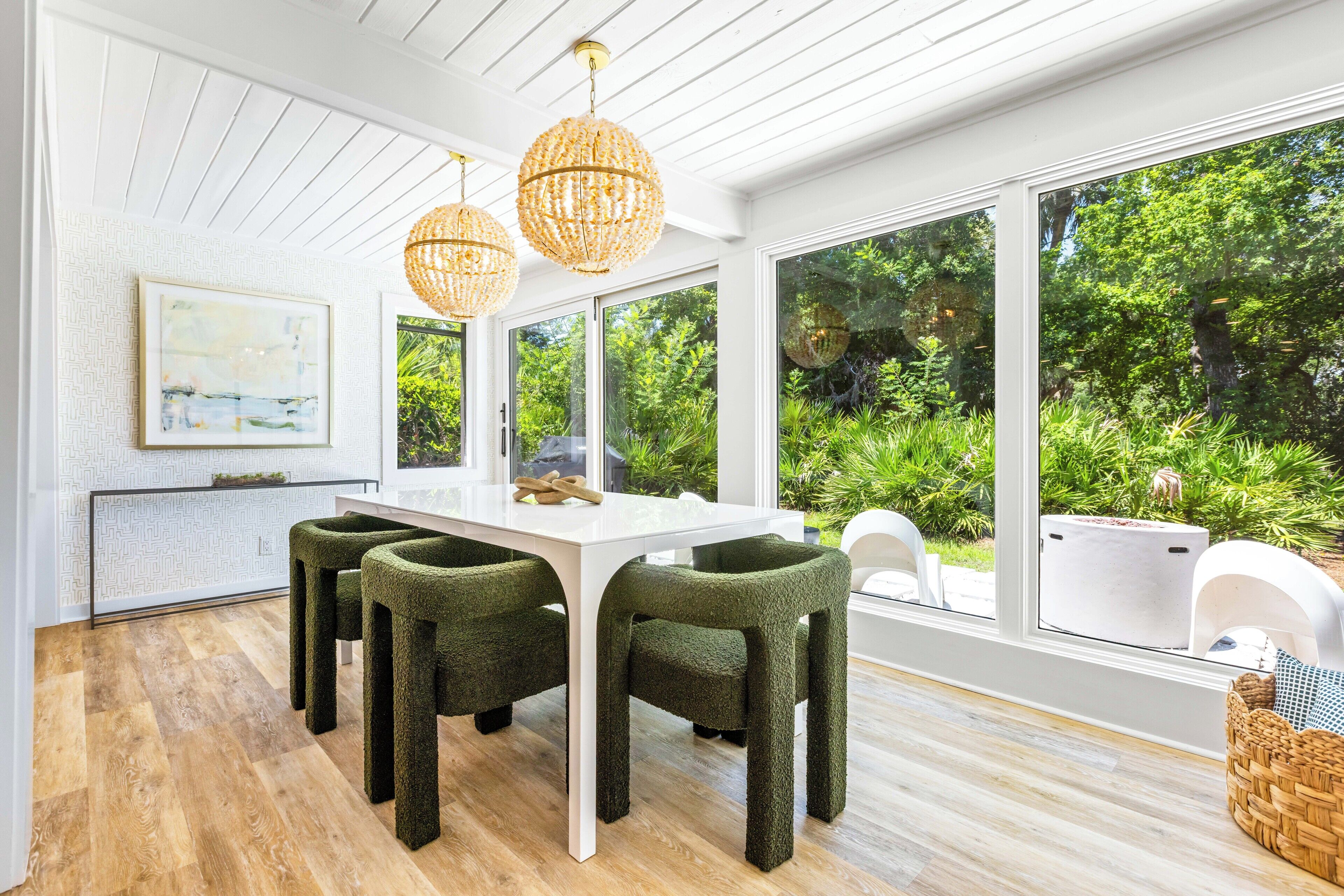 A modern dining area with a white table and green upholstered chairs, large windows offering a view of lush greenery, and a hanging light fixture.
