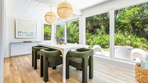 Un moderno comedor con una mesa blanca y sillas tapizadas en verde, ventanas amplias que ofrecen vistas a exuberante follaje y una lámpara colgante.