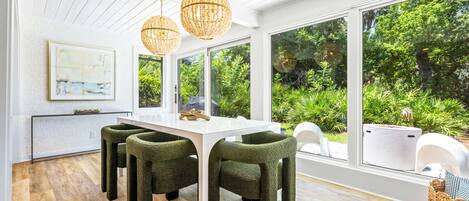 A modern dining area with a white table and green upholstered chairs, large windows offering a view of lush greenery, and a hanging light fixture.