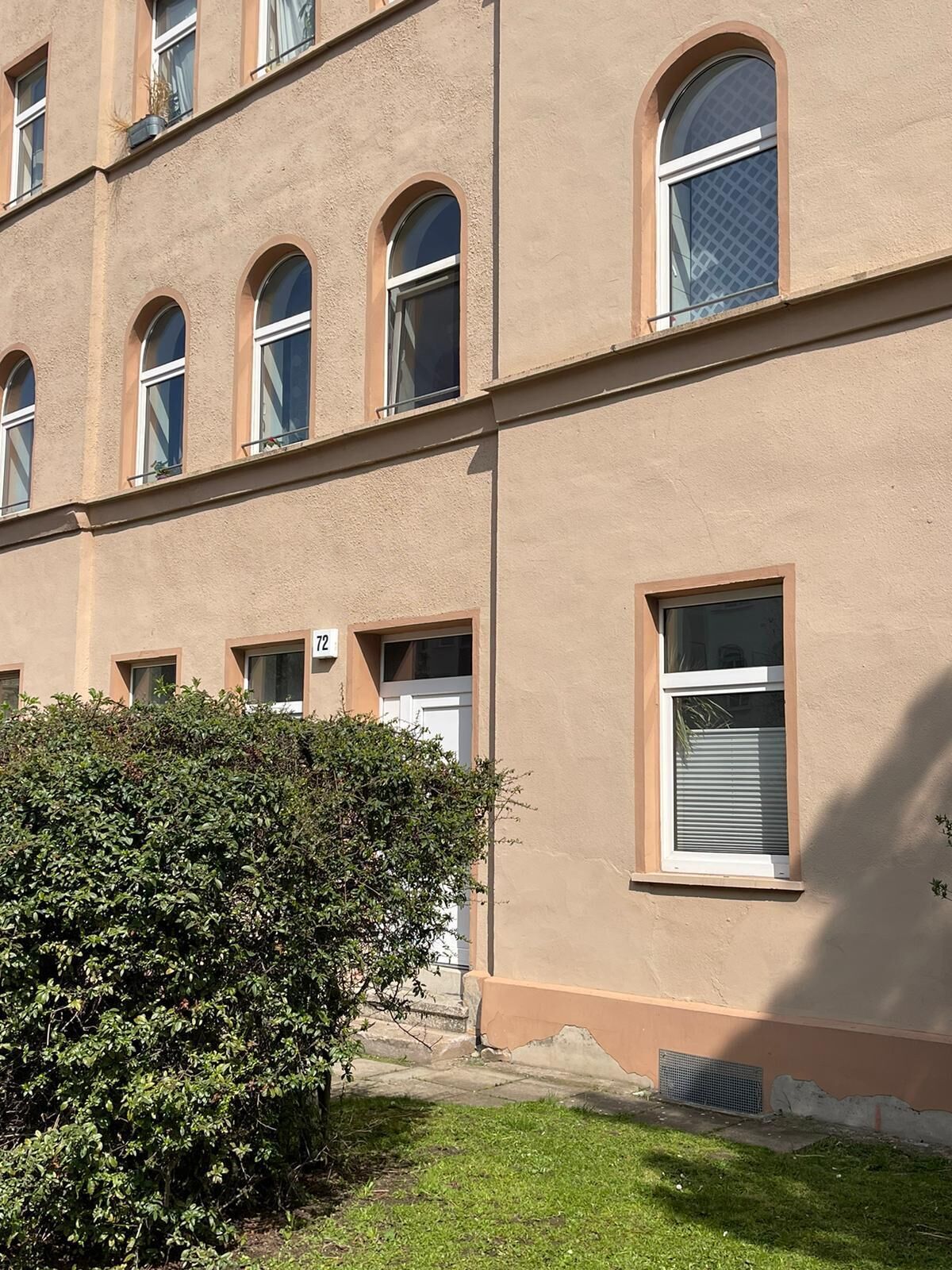 Ein mehrstöckiges Gebäude mit einer beigefarbenen Fassade, zahlreichen Fenstern und einer kleinen Grünfläche davor.