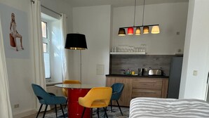 Et moderne soveværelse med en stor seng, et spiseområde med et rødt bord og blå stole, et køkken med et køleskab, og en pendel over spisebordet.