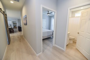 A hallway leading to a bedroom with a ceiling fan, a bathroom with a toilet, and a kitchen area with cabinets and a countertop.