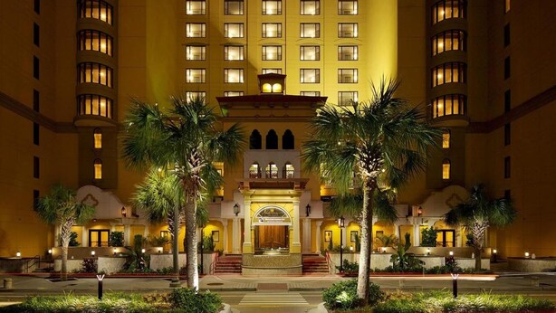 Un hotel de varios pisos con entrada central, palmeras y ventanas iluminadas.