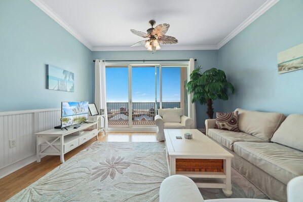 A spacious living room with a large window, a ceiling fan, a white sofa, a coffee table, and a TV. - Top-Floor 2BR/2BA Condo with Stunning Gulf Views | Sleeps 6 (Gulf Shores)