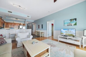A spacious living room with a white sofa, a coffee table, and a TV on a white stand. There is a kitchen area with wooden cabinets and a dining table with chairs. - Top-Floor 2BR/2BA Condo with Stunning Gulf Views | Sleeps 6 (Gulf Shores)