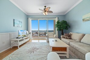 Un salon spacieux avec une grande fenêtre, un ventilateur de plafond, un canapé blanc, une table basse et un téléviseur.