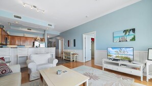 Un amplio salón con un sofá blanco, una mesa de centro y un televisor sobre un mueble blanco. Hay una zona de cocina con armarios de madera y una mesa de comedor con sillas.