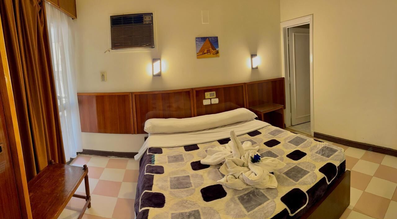 Hotellihuone, jossa on sänky, puinen penkki, ruutukuviollinen lattia, seinällä oleva taulu ja ovi.