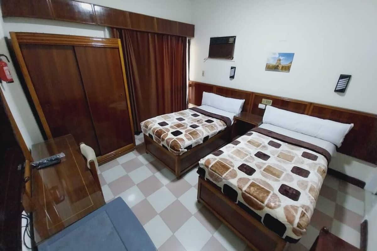Quarto de hotel com duas camas, um guarda-roupa de madeira, uma escrivaninha e piso xadrez.