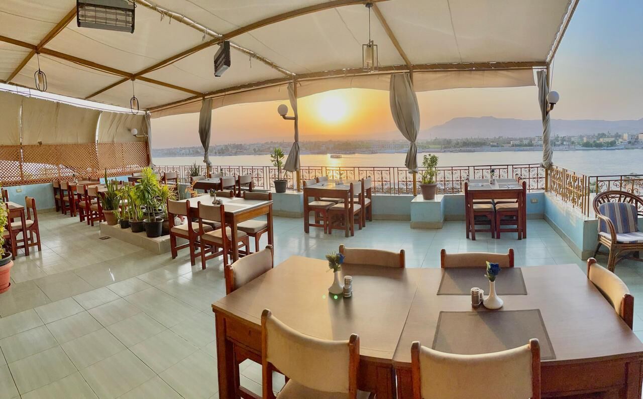 Un espace extérieur abrité, aménagé pour les repas, avec des tables et des chaises, des plantes en pot, et une vue sur un plan d’eau et des montagnes au coucher du soleil.