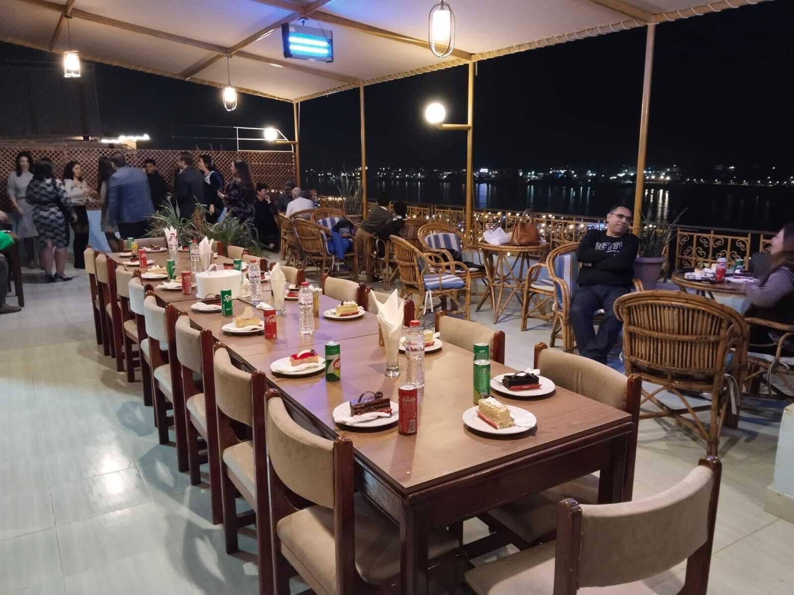 Un espacio de comedor al aire libre con mesas y sillas de madera, rodeado por una barandilla y vista nocturna de la ciudad.