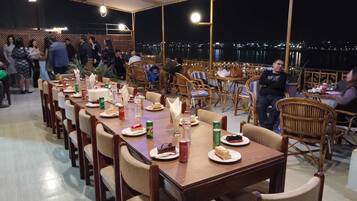 Un espacio de comedor al aire libre con mesas y sillas de madera, rodeado por una barandilla y vista nocturna de la ciudad.