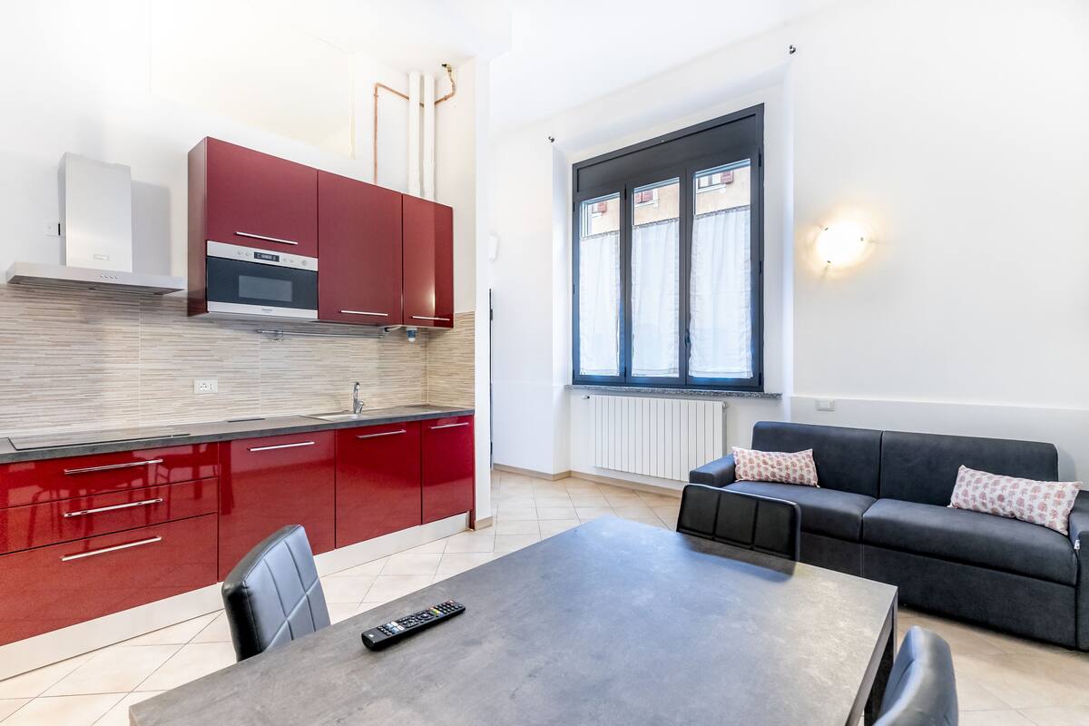 A modern kitchen with red cabinets, a dining area with a table and chairs, and a living area with a sofa and a window with curtains.