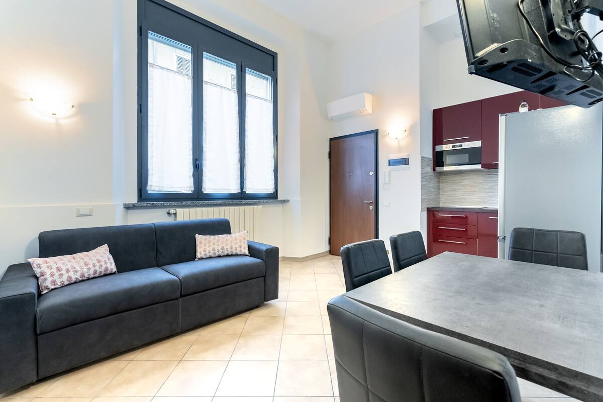 A modern living room with a dark sofa, a dining table, and a kitchen area with red cabinets and a white refrigerator.