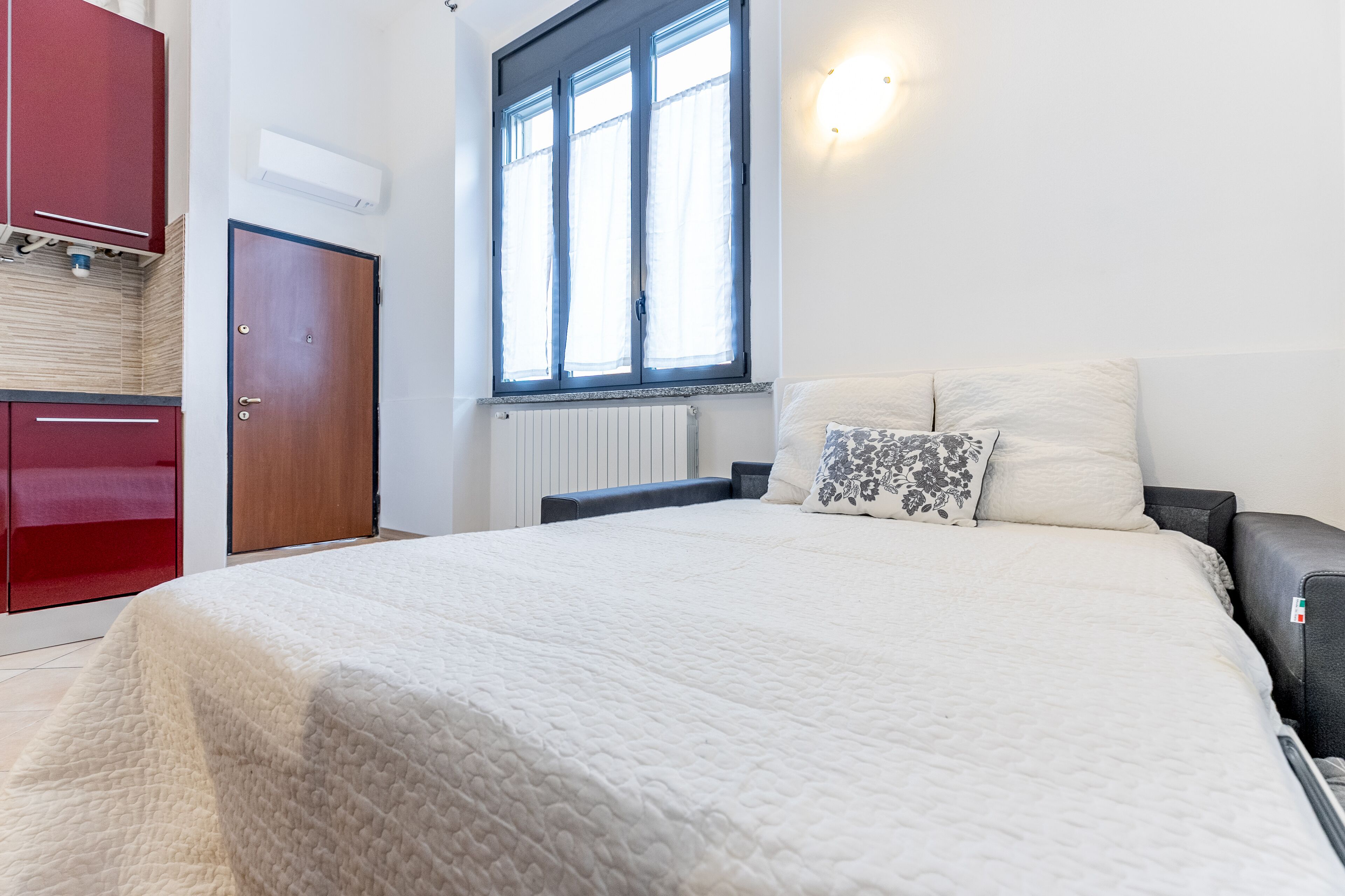 A bedroom with a large bed, a wardrobe, a window with curtains, and a wall-mounted light fixture.