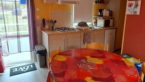 A compact kitchen with a dining area, featuring a table with a red and yellow tablecloth, wooden chairs, and a view of the outside through a window. - Mobil Home à 1.5km du Centre Ville, en en Plein Cœur de la Montagne Corse (Corte)