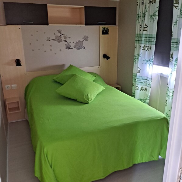 Une chambre avec un lit simple, une couvre-lit verte, une tête de lit ornée de motifs floraux et une fenêtre avec des rideaux à motifs de bambou.