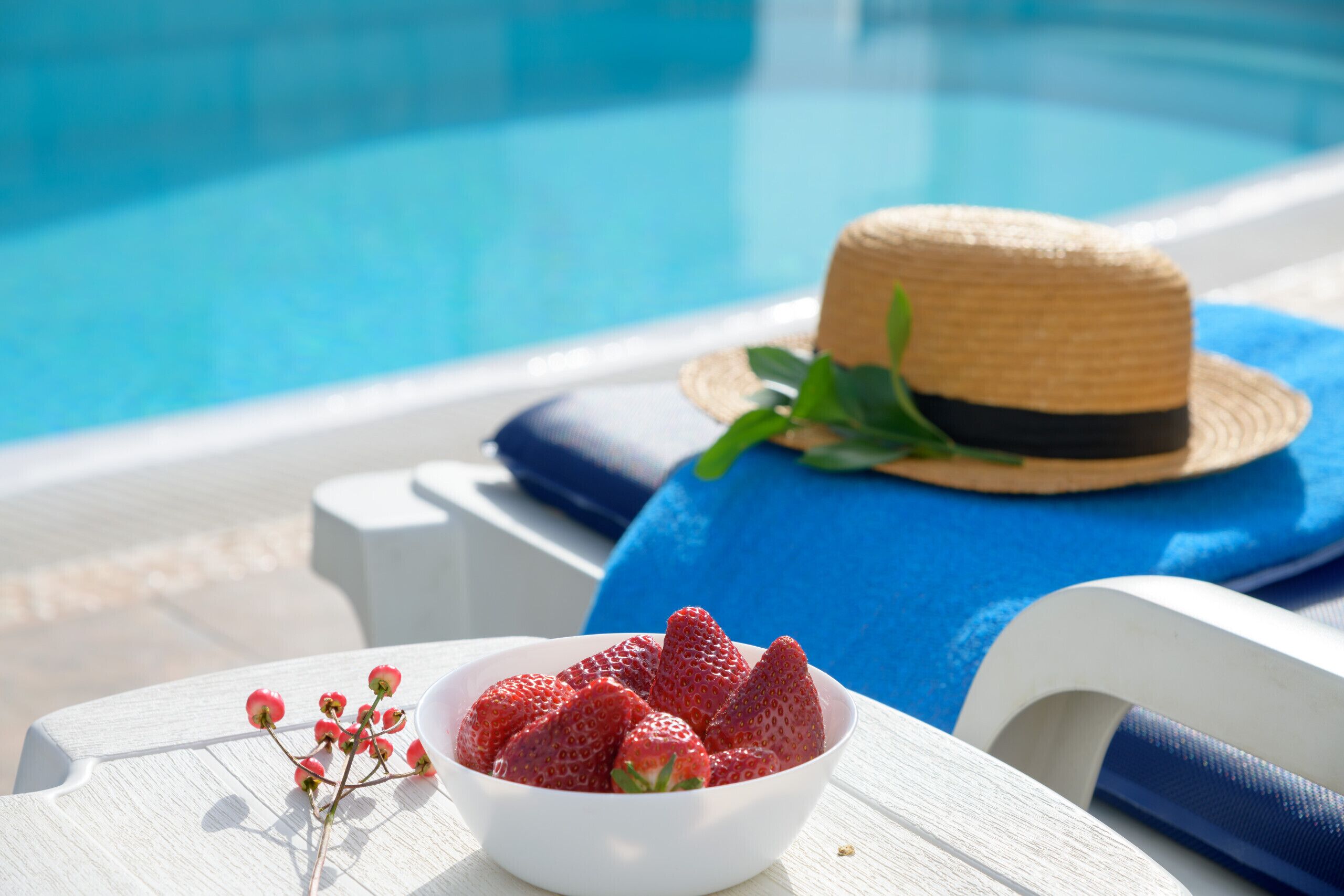 A poolside setting with a white lounge chair, a straw hat on a blue towel, and a bowl of strawberries.