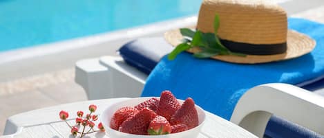 Escena junto a la piscina con una silla de descanso blanca, un sombrero de paja sobre una toalla azul y un bol de fresas.