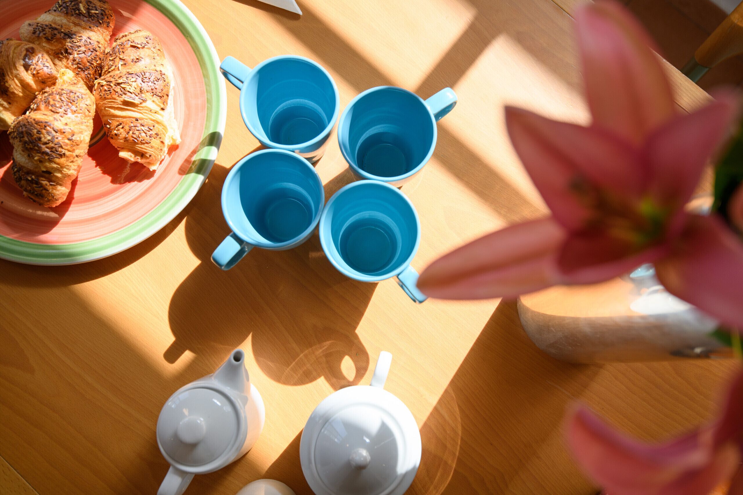 Un tavolo in legno con croissant, tazze blu, teiere bianche e un fiore rosa.