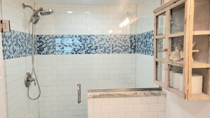 Un baño con azulejos blancos, una ducha con cabezal de ducha manual y un ventanal con marco de madera.
