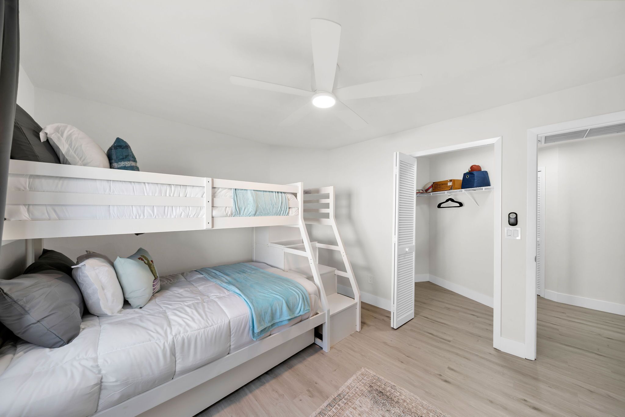 Une chambre avec un lit superposé, un ventilateur de plafond, un petit bureau et une porte donnant sur une autre pièce.
