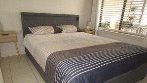 A neatly made bed with striped bedding, a wooden nightstand with a lamp, and a window with blinds.