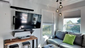 Un salon moderne avec un téléviseur à écran plat fixé au mur, un bar en bois avec des tabourets, un canapé gris agrémenté d’un coussin vert et une grande fenêtre avec des stores.