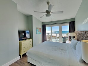 Une chambre avec un lit, un téléviseur posé sur une commode, un ventilateur de plafond et une vue sur la plage, visible à travers une porte coulissante.