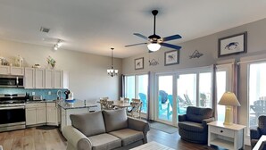 A spacious living area with a sofa, coffee table, and a dining table. There is a kitchen with modern appliances and a ceiling fan. The room has large windows with a view of the outdoors. - Charline Beachside Villa | Gulf Front Luxury (Panama City Beach)