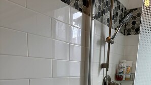A bathroom with white tiled walls, a shower with a handheld showerhead, and a towel rack with folded towels.
