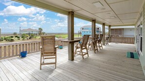 Eine überdachte Terrasse im Freien mit Holzsesseln und -tischen mit Blick auf einen Strand und Häuser.