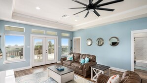 Ein geräumiges Wohnzimmer mit Deckenventilator, einem braunen Ledersofa, einem Couchtisch und Blick durch große Glastüren auf eine Außenpatio.