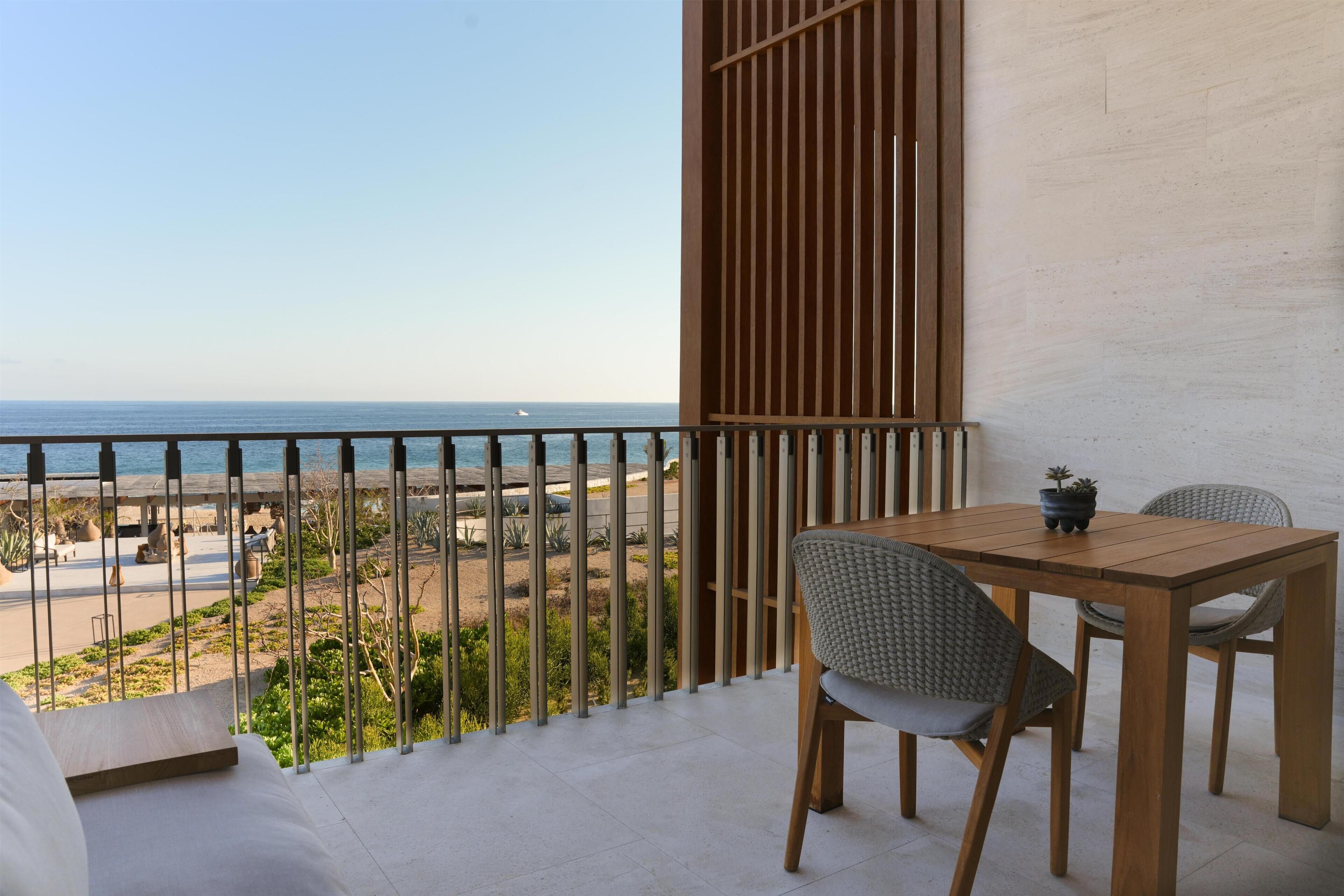 A balcony with a view of the beach and ocean, a wooden table with two chairs, and a potted plant.