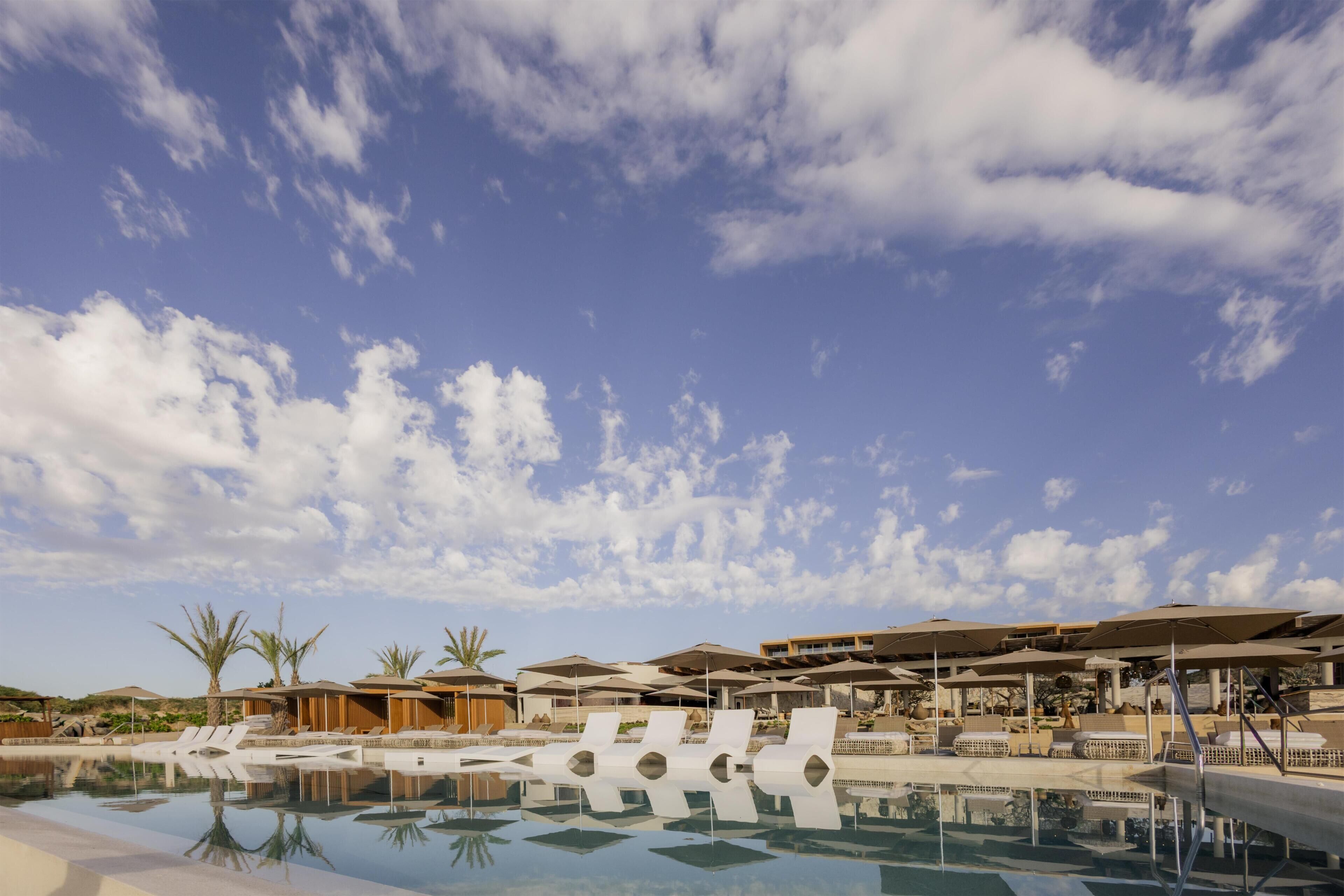 A resort pool area with lounge chairs and umbrellas, surrounded by buildings and palm trees.