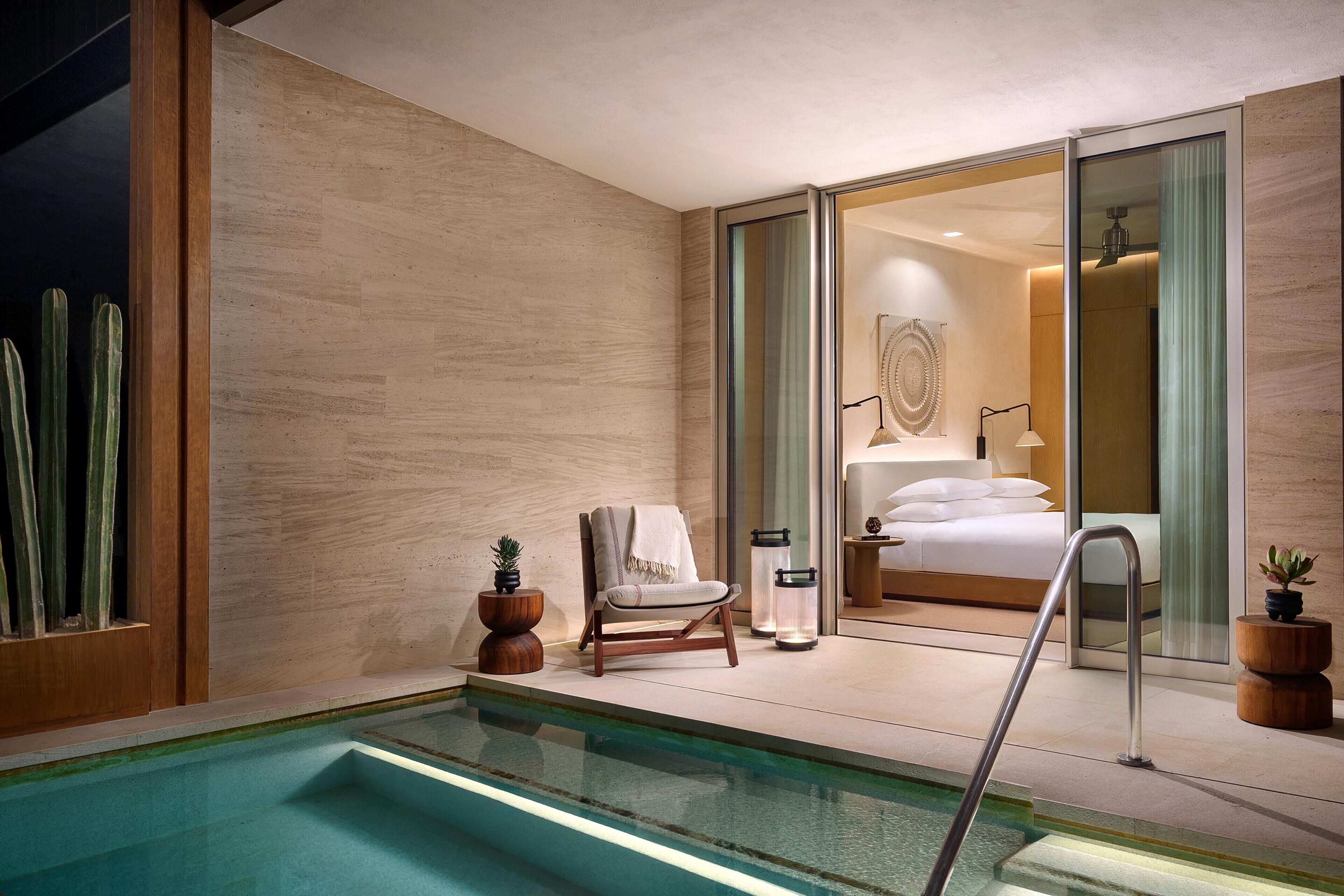 A modern hotel room with a glass door leading to a bedroom, a small pool, and a seating area with a chair and plants.