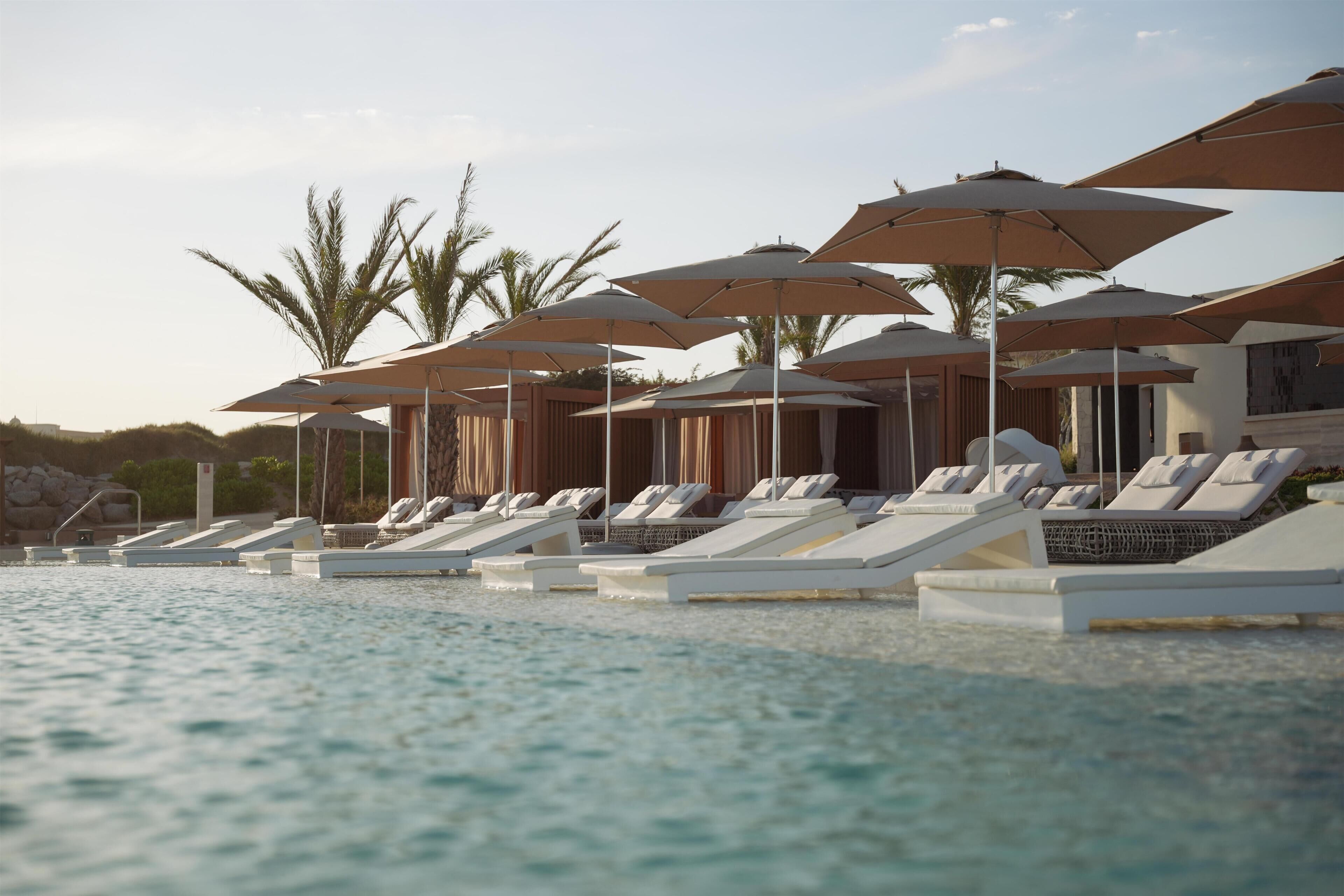 Zona junto a la piscina con sillones de descanso y sombrillas, rodeada de palmeras.