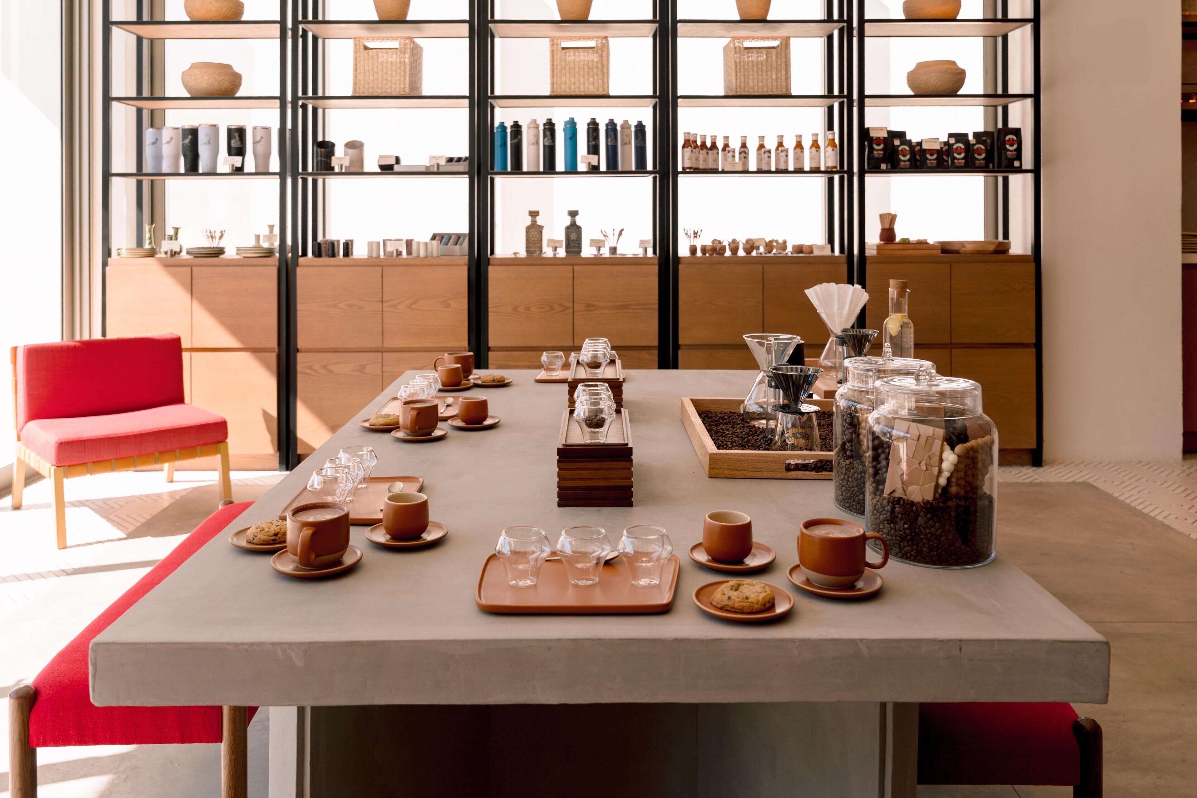A café setting with a long table set for coffee service, a red chair, and shelves stocked with various items in the background.