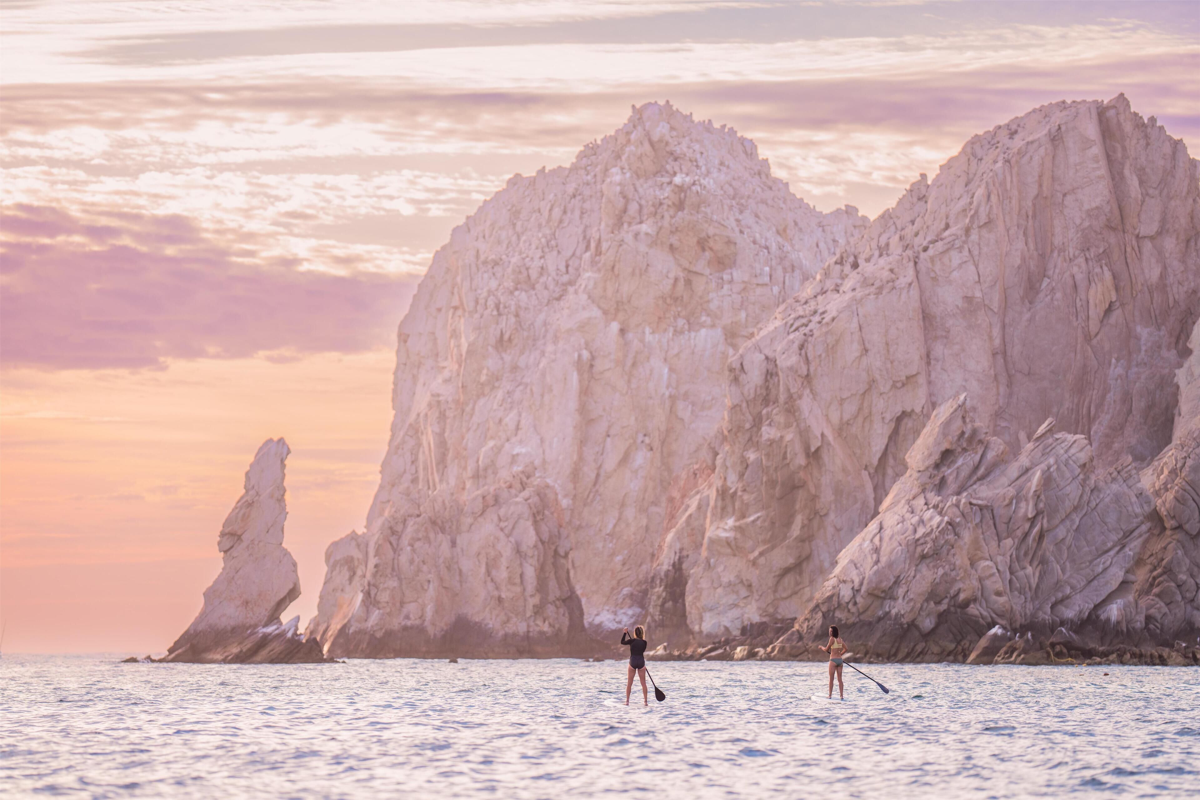 A coastal scene with two individuals paddleboarding near a rocky cliff.