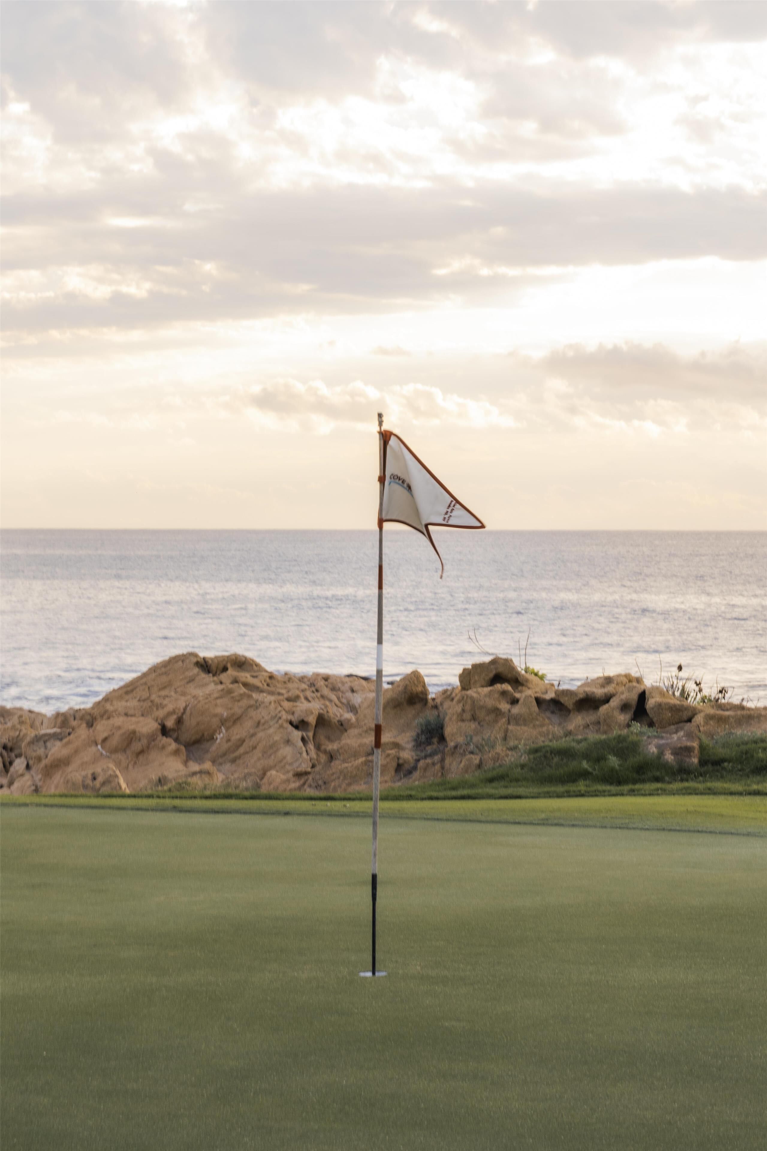 A golf course with a flagstick in the foreground and a rocky shoreline in the background.
