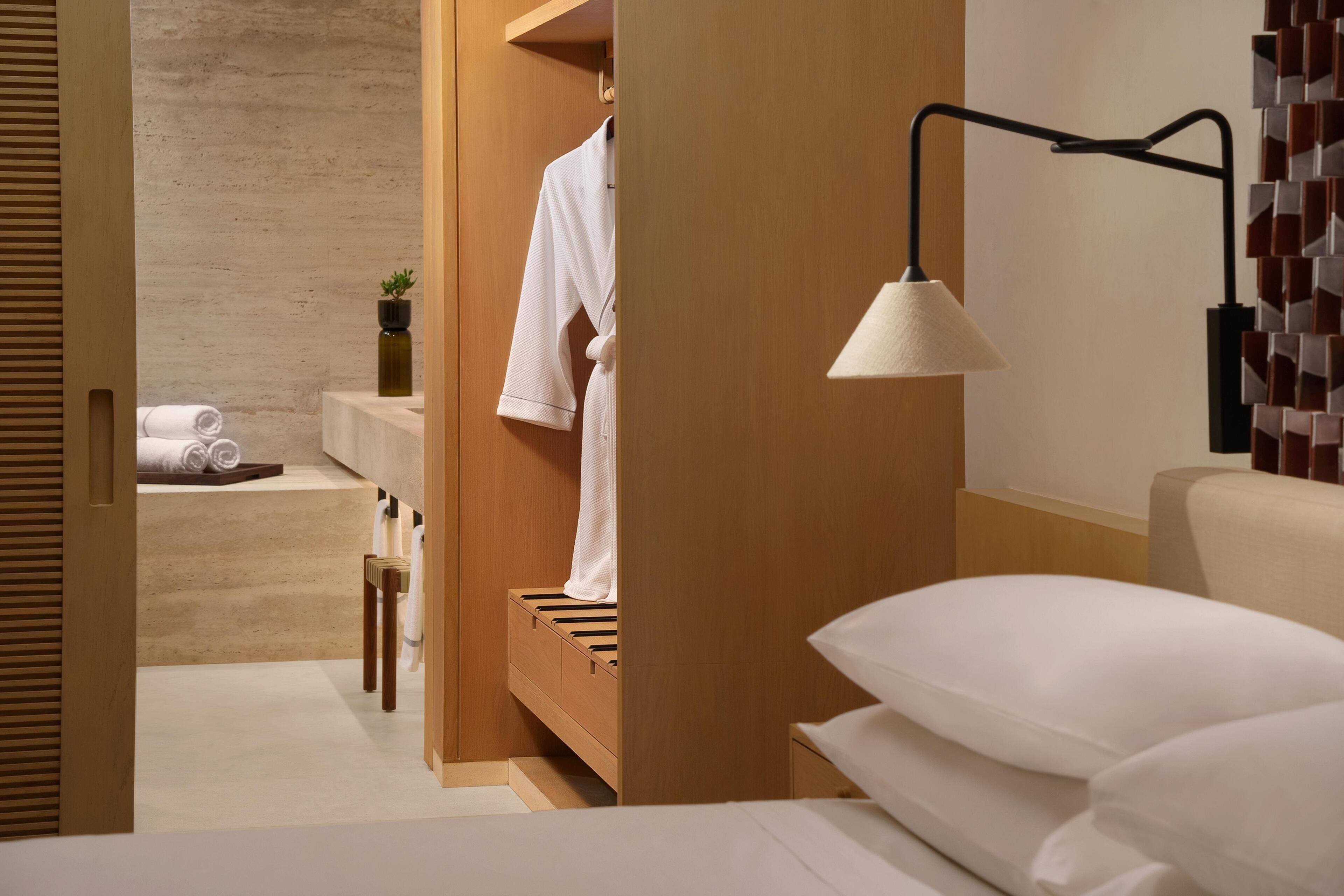A hotel room with a wooden wardrobe, a bedside table, a lamp, and a bed with white linens.