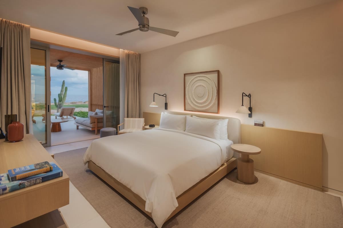 Een moderne hotelkamer met een groot bed, een plafondventilator, uitzicht op het strand en een kleine tafel met een lamp.