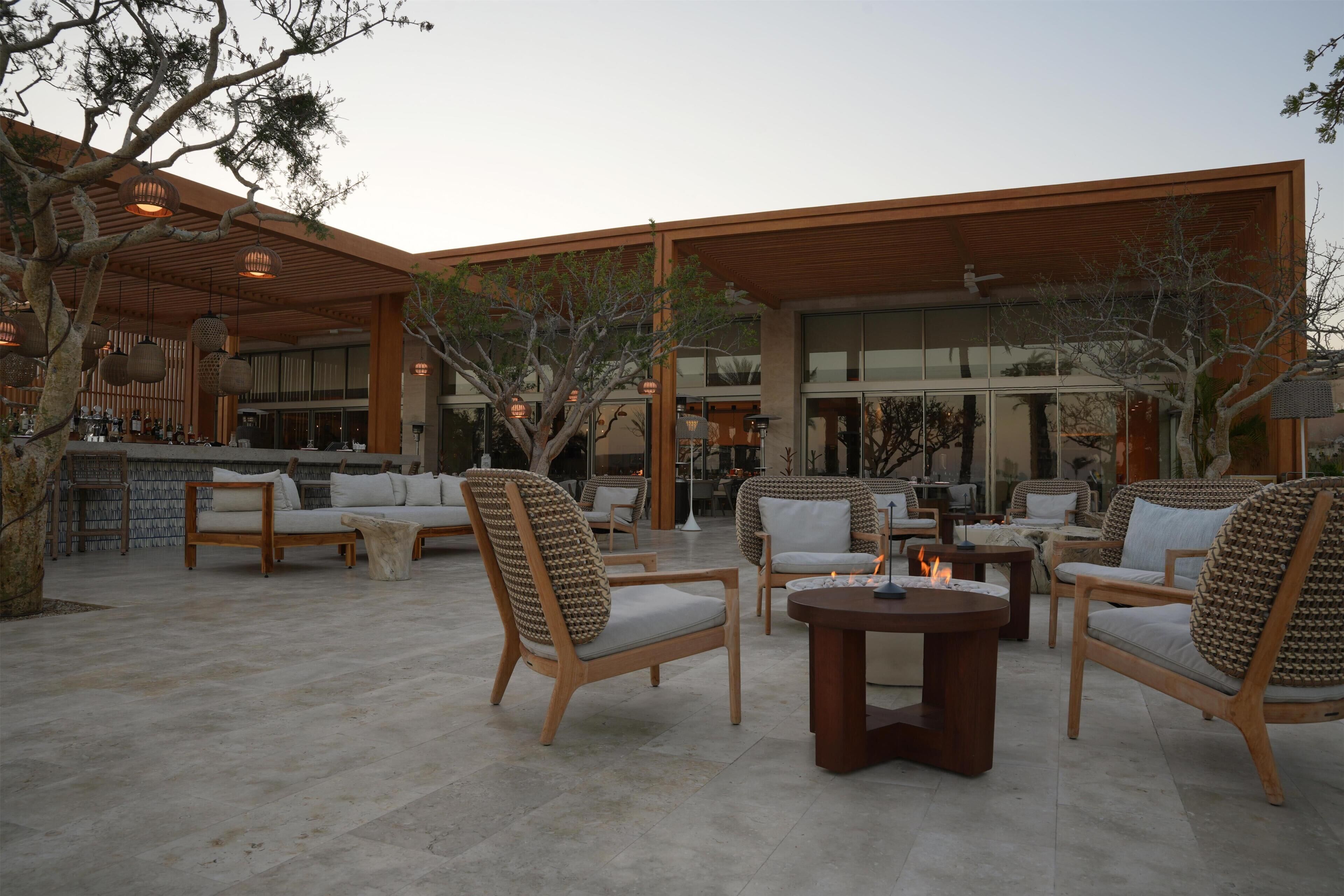 An outdoor seating area with wicker chairs and a fire table, surrounded by a modern building with large windows.