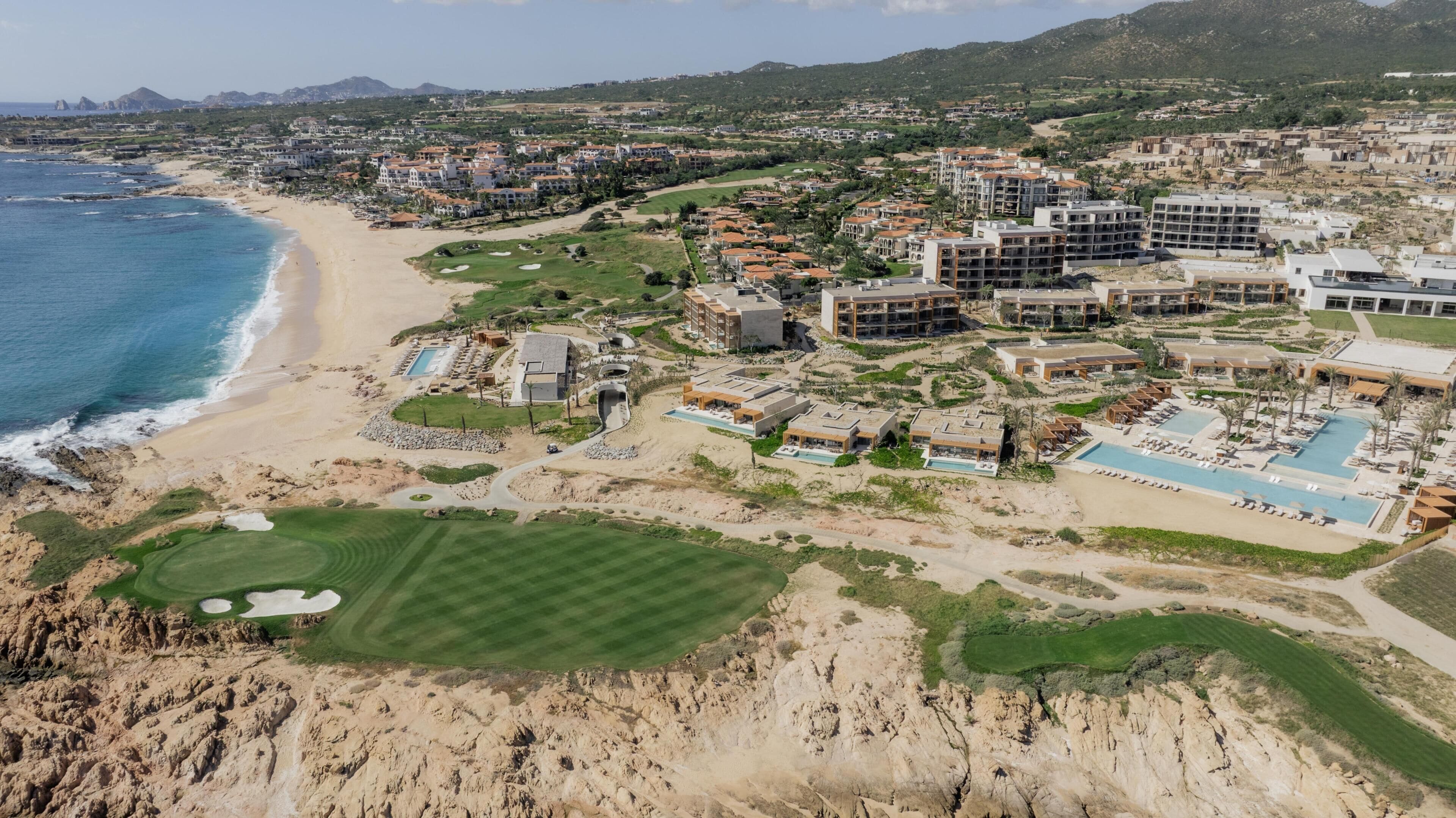 Vista aérea de un complejo turístico costero con varios edificios, una playa y un campo de golf.